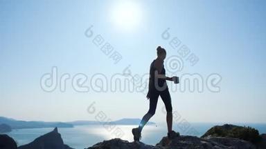 一位快乐的徒步旅行者女士的剪影，她端着一杯茶站在山顶，从高处欣赏着美妙的景色